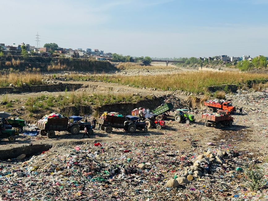 A small vintage pick-up truck painted in faded blue with red rimmed wheels is parked parallel to the roadside. Its cargo area is loaded with a large assortment of various waste materials including fla